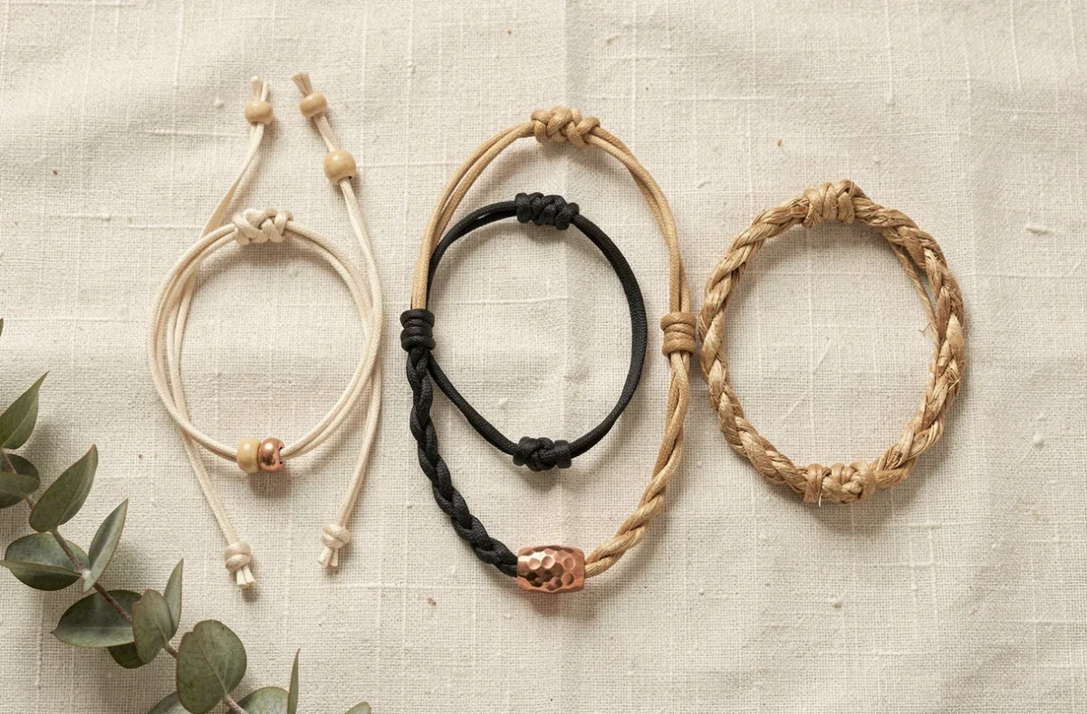 Flat lay de cinq bracelets cordon minimalistes variés sur tissu lin blanc cassé avec détails nœuds et fermoirs